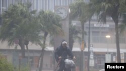 জাপানের দ্বীপ কিউশু-তে টাইফুন নানমাডলের কারণে সৃষ্ট প্রবল বৃষ্টি ও বাতাসের মধ্যে সাইকেলে চড়ে একজন ব্যক্তি যাতায়াত করছেন। ১৮ সেপ্টেম্বর, ২০২২। (রয়টার্সের মাধ্যমে কিয়োডো)